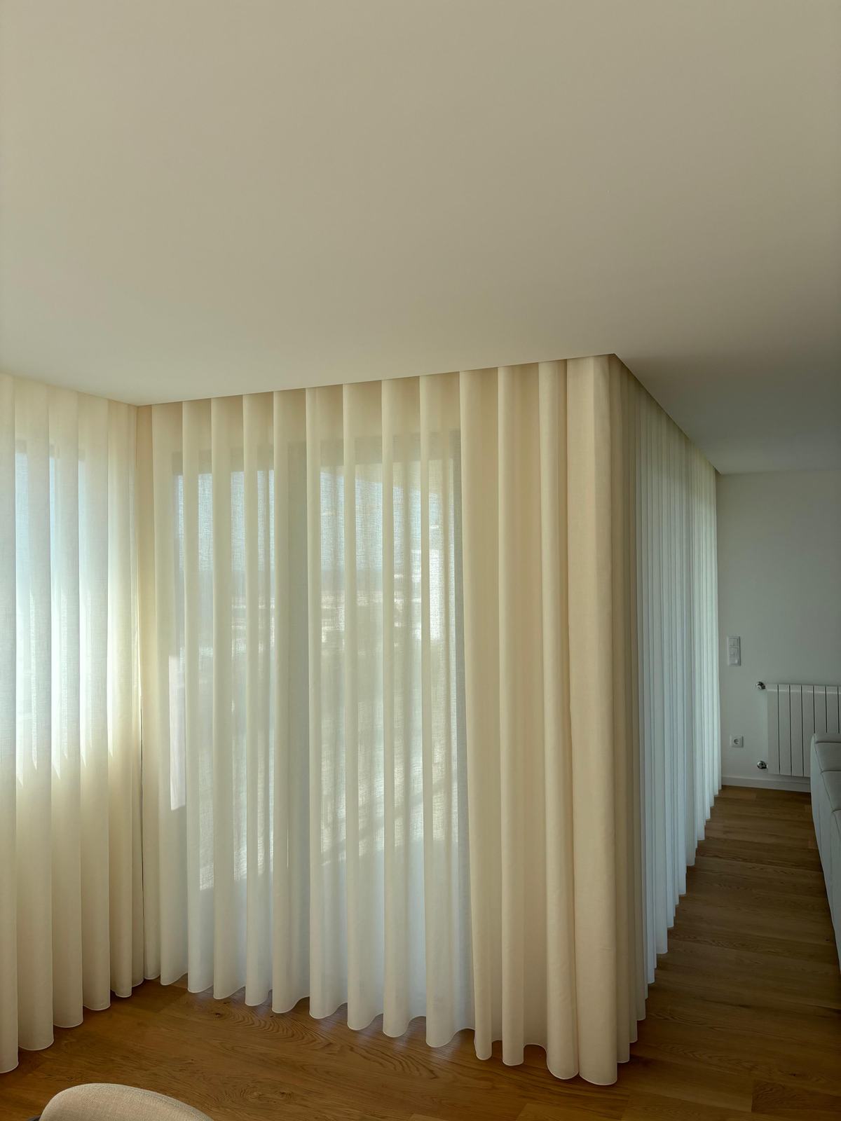 Cortinado de onda em tom creme a dobrar o canto de uma divisão com chão de madeira