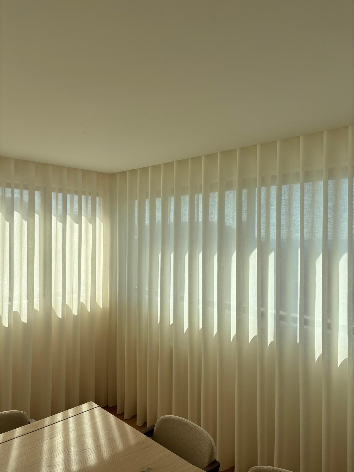 Cortinados de onda em tom creme numa sala de jantar, com o sol a atravessar o tecido