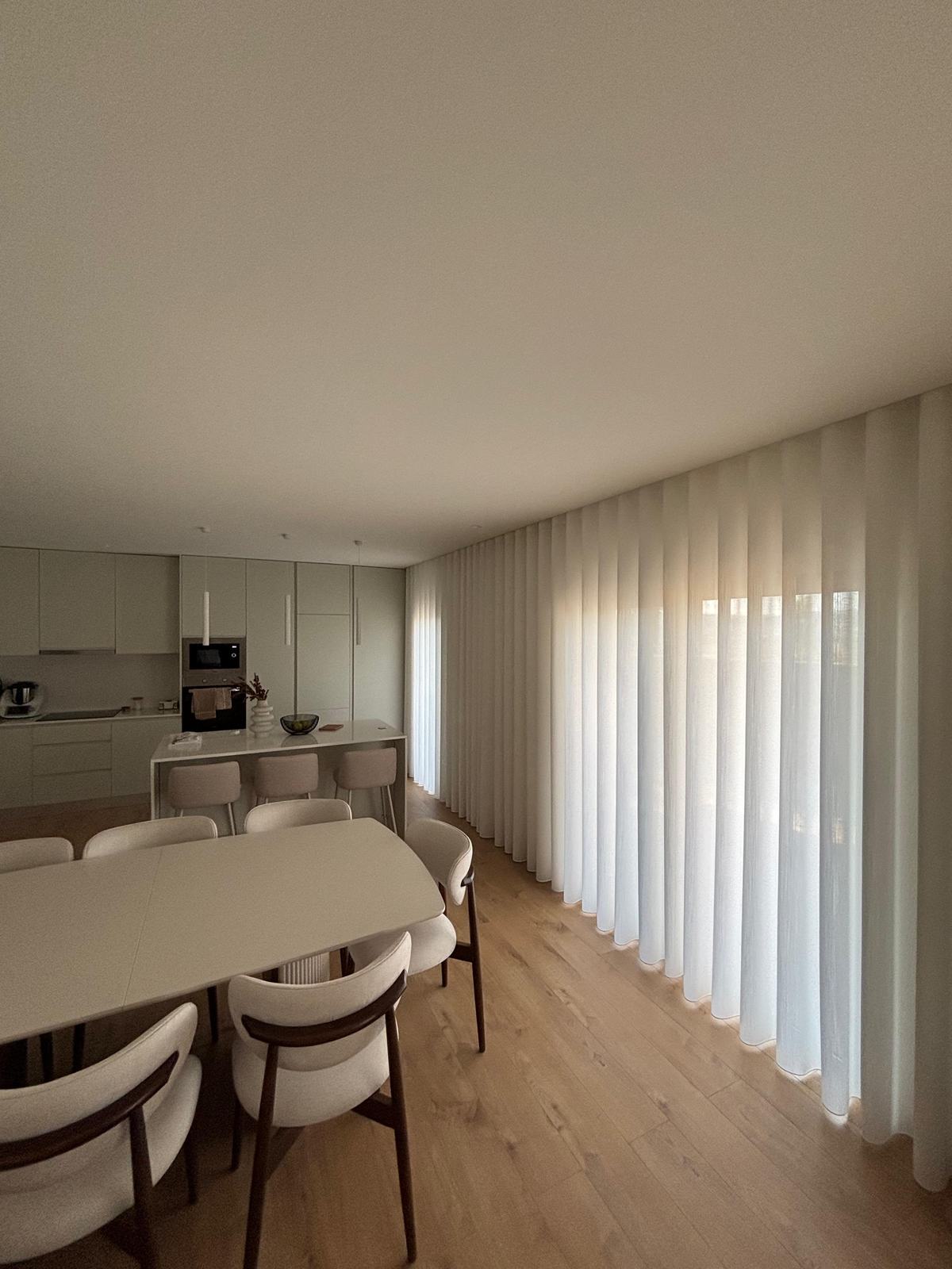 Cozinha e sala de jantar em open space com cortinados de onda claros ao longo do envidraçado