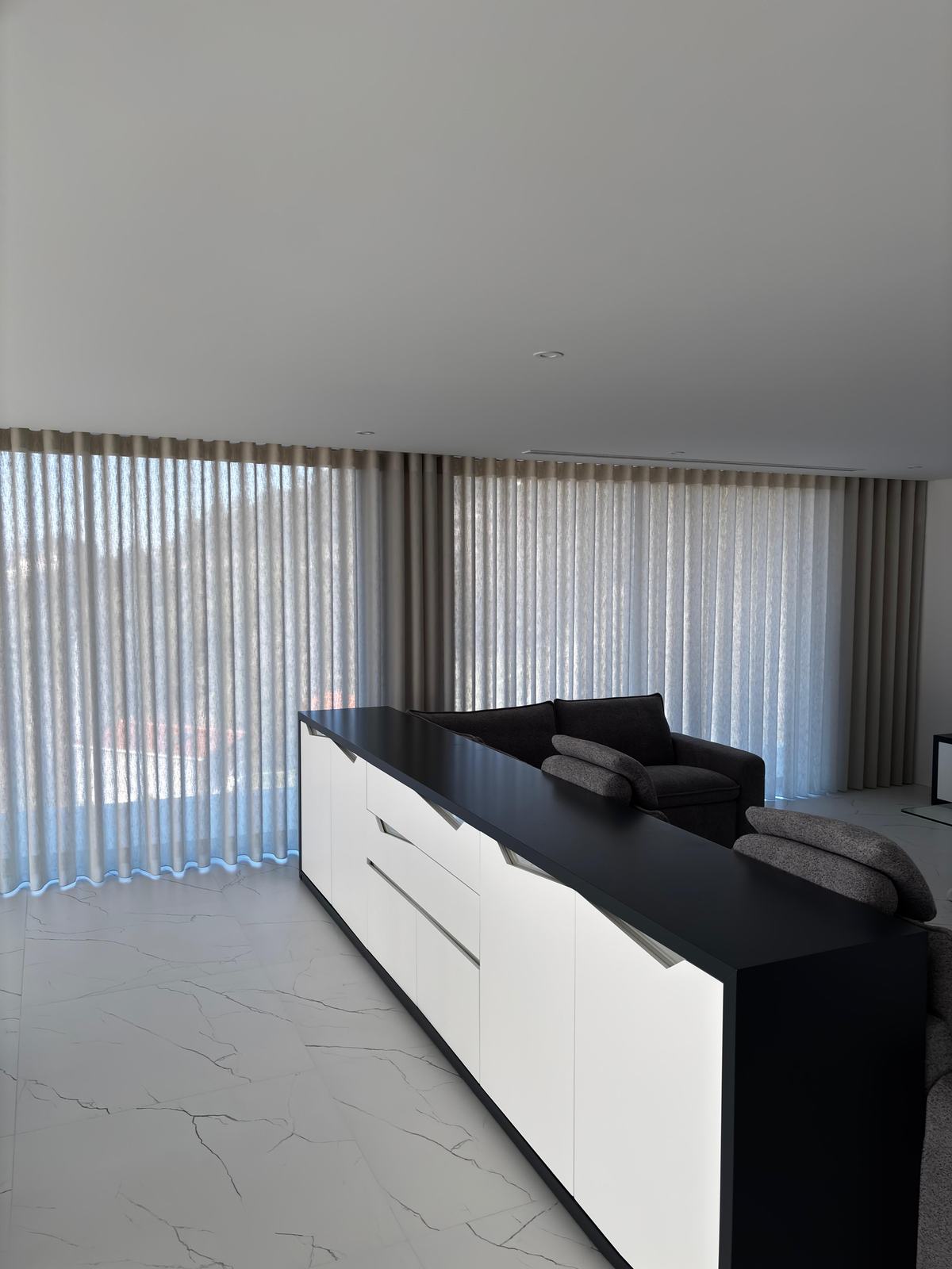 Sala aberta com um móvel branco e preto e cortinados de onda translúcidos em dois envidraçados