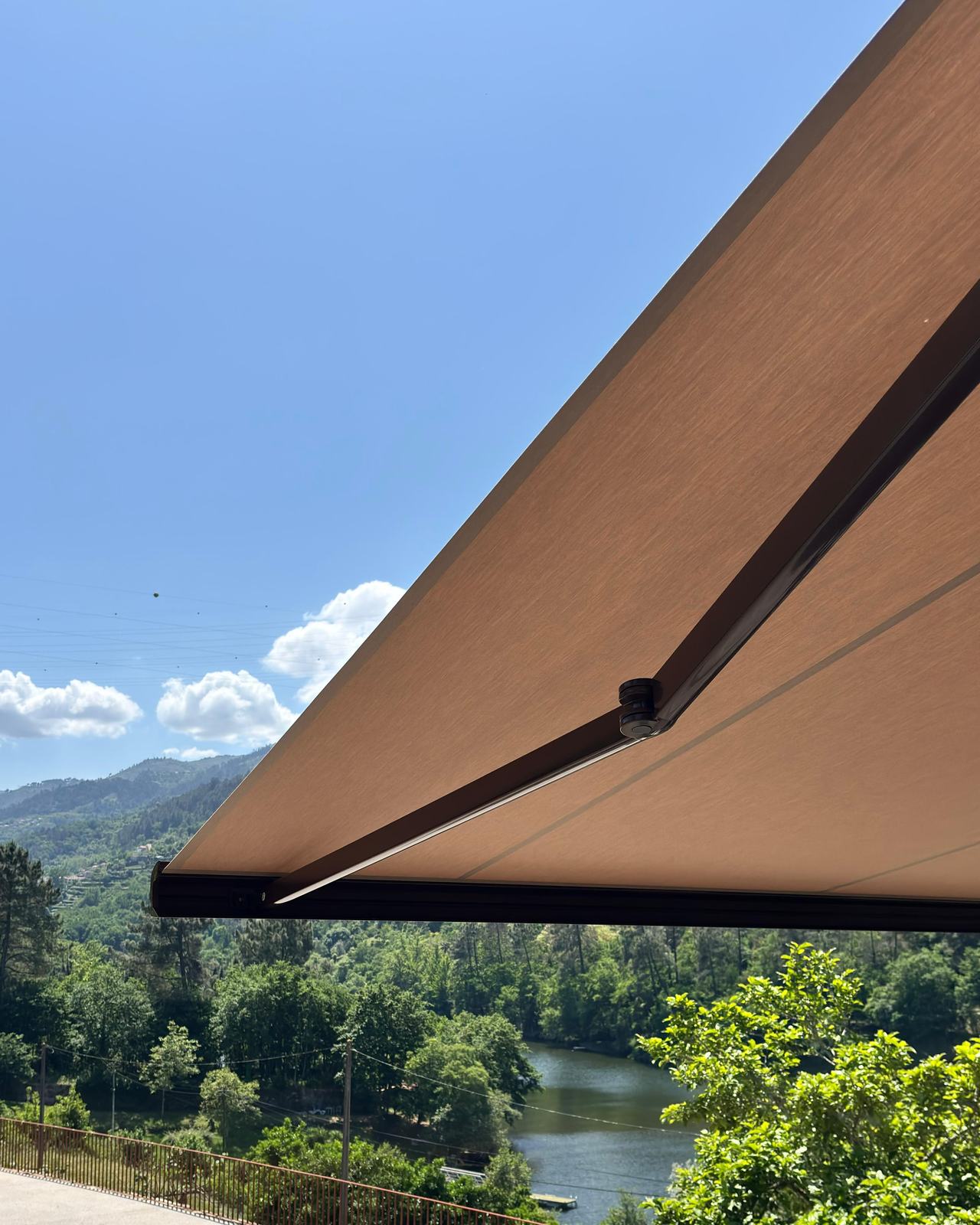 Pormenor do braço articulado e da tela de um toldo aberto, com o rio e a serra ao fundo